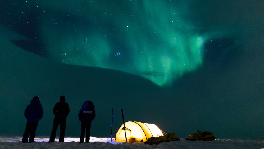 Trois personnes regardent une aurore boréale à côté de leur tente